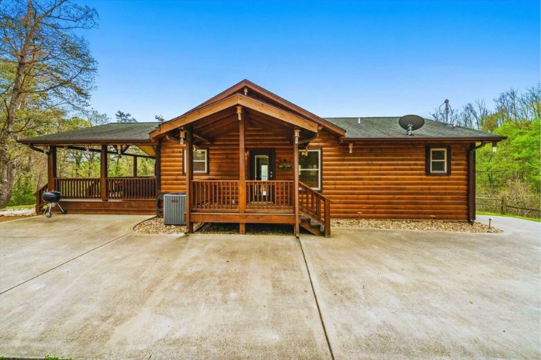 Single-story log cabin with a covered front porch and wraparound deck, set on a wide concrete driveway and surrounded by trees under a clear blue sky.